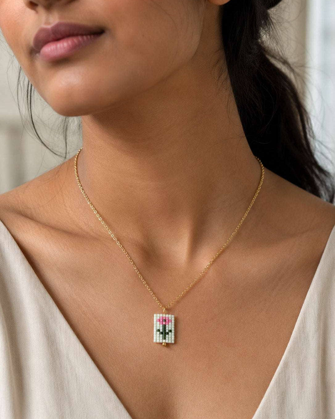 A close-up lifestyle shot of a woman wearing a delicate gold chain necklace with a small rectangular pendant featuring a colorful pixel-art style design.