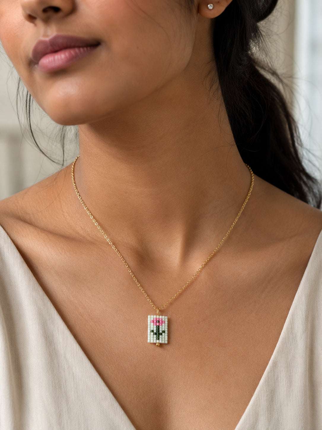 A close-up lifestyle shot of a woman wearing a delicate gold chain necklace with a small rectangular pendant featuring a colorful pixel-art style design.